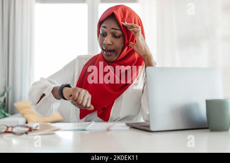 Donna d'affari musulmana afro-americana seduta alla scrivania di fronte al portatile, guardando l'orologio e toccando la testa Foto Stock