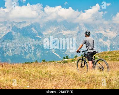 L'adolescente si siede sul moderno mtb e guarda dalla cima della collina a cime stonny e nevoso di fronte. Alpi dolomitiche, regione di Trento, Italia Foto Stock