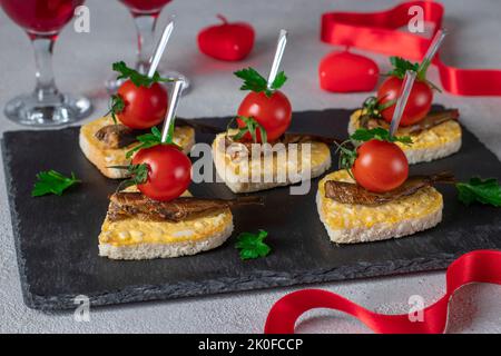 Canape a forma di cuore con spratti, formaggio, uovo e pomodori ciliegini su piatto di ardesia su sfondo grigio chiaro, idea per San Valentino Foto Stock