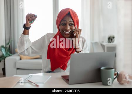 Nuove opportunità di lavoro. Donna musulmana afro-americana molto gioiosa che parla sul cellulare e celebra il successo Foto Stock