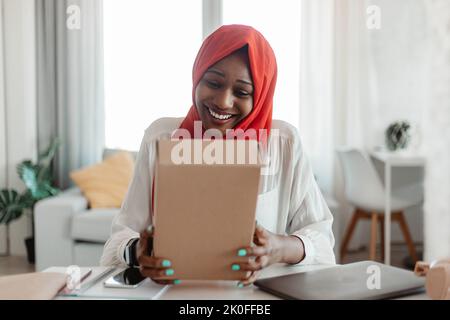 Ragazza musulmana afro-americana eccitata che tiene la scatola di cartone e sorridente, facendo l'ordine su Internet con la consegna Foto Stock