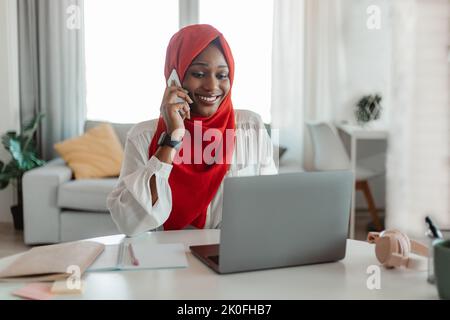 Donna afro-americana felice in hijab che lavora sul portatile in ufficio domestico e che parla su smartphone con il cliente Foto Stock