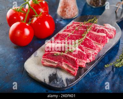 Su sfondo blu scuro, un tagliere con bistecche di carne cruda con un rametto di rosmarino, appetitosi pomodori maturi e spezie. Primo piano. Cucinare carne Foto Stock