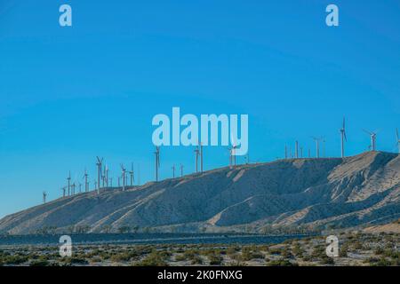 Mulini a vento sulla cima di una montagna in un deserto in California. Vista delle turbine eoliche dalla terra desertica con piante selvagge e vista di un cielo azzurro chiaro. Foto Stock
