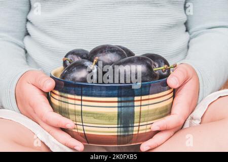 Il bambino tiene le prugne in una ciotola di ceramica. Frutta raccolta dal proprio giardino alla stagione di raccolta. Frutta matura fresca e succosa. Particolare delle mani del ragazzo. Foto Stock