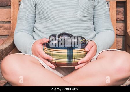 Bambino che detiene raccolto fresco di prugne biologiche. Succosa casa coltivata. Frutta matura in ciotola di ceramica nelle mani dei bambini. Dal mercato locale. Ragazzo che aiuta in giardino. Foto Stock