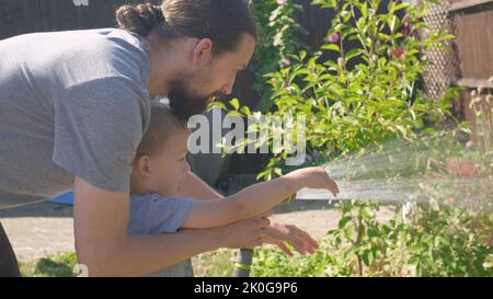 Padre e Figlio. Divertente bambino annaffiatura piante prato in giardino casa cortile con papà. Lavori di casa estivi. Sgabello duro Kid all'aperto Foto Stock