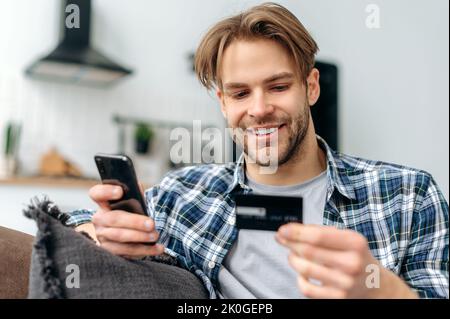 Positivo soddisfatto ragazzo caucasico sorridente, in abiti casual elegante, si siede sul divano a casa, tiene in mano il telefono cellulare e la carta di credito, paga per gli acquisti online, inserisce un numero di carta, shopping online Foto Stock