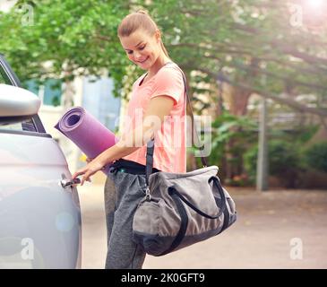 Sulla sua strada per il fitness. Una bella giovane donna che porta un tappetino yoga mentre entra in auto. Foto Stock