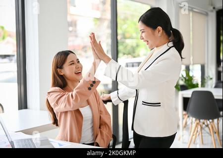 Due giovani donne d'affari asiatiche felici e allegre stanno ridendo e celebrando il loro successo insieme, dando i high-five in ufficio. Concep. Lavoro di squadra Foto Stock