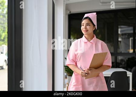 Bella giovane e affascinante infermiera asiatica in uniforme con appunti medici si trova nel corridoio dell'ospedale. Concetto di personale sanitario Foto Stock