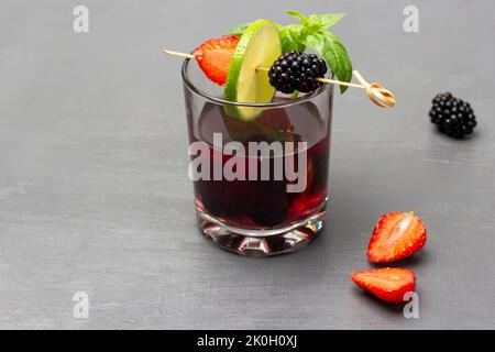Bevanda estiva infusa con more e basilico verde in vetro. Lime, fragole e mora sullo spiedino. Spazio di copia. Sfondo grigio Foto Stock