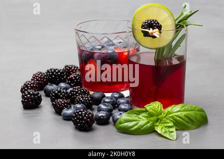 Due bicchieri di bevanda ai frutti di bosco. BlackBerry e lime cuneo su spiedino. More, mirtilli, foglie di basilico verde sul tavolo. Primo piano. Sfondo grigio. Foto Stock