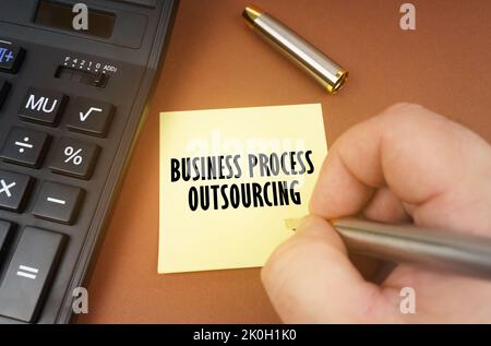 Concetto aziendale. Una calcolatrice si trova su una superficie marrone, una mano con una penna fa un'iscrizione su un adesivo - Business Process Outsourcing Foto Stock