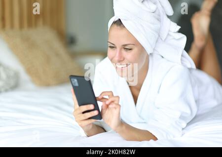 Donna felice dopo aver fatto la doccia utilizzando il telefono cellulare sdraiato sul letto a casa Foto Stock