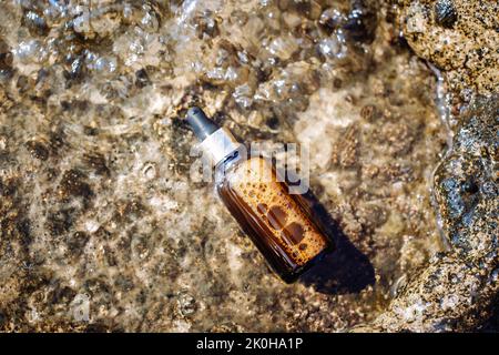 Prodotto cosmetico in mare su una pietra. Bottiglia di siero di vetro marrone in acqua su una riva rocciosa. Vista dall'alto, disposizione piatta. Foto Stock