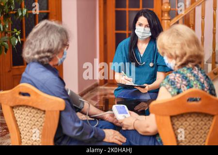 Il medico che usa lo sfigmomanometro assume la pressione sanguigna di pazienti anziani durante la visita a casa Foto Stock