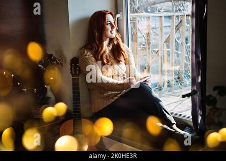 Interni accoglienti Hobby per l'inverno, stagione fredda autunno. Donna rossa che suona la chitarra acustica e ascolta la musica, seduta vicino alla finestra di casa Foto Stock