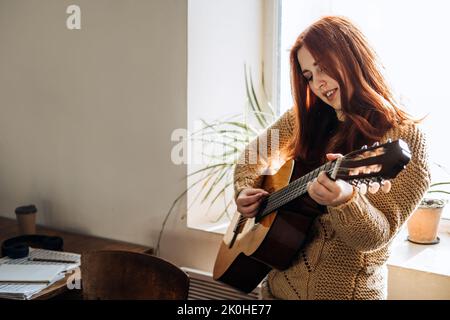 Interni accoglienti Hobby per l'inverno, stagione fredda autunno. Donna rossa che suona la chitarra acustica e ascolta la musica, seduta vicino alla finestra di casa Foto Stock