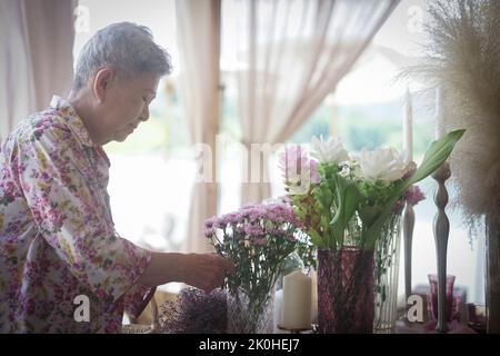 vecchia donna decorare bouquet di fiori in vaso di vetro sul tavolo da pranzo Foto Stock