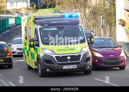 Primo piano vista frontale di Fiat NHS luce blu ambulanza di emergenza sorpasso coda di traffico seguita da auto di polizia a Billericay High Street Inghilterra Regno Unito Foto Stock