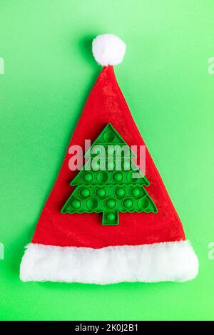Immagine di Natale con trendy popolare bambino anti-stress pop giocattolo in forma di verde albero di Natale sul cappello rosso di Babbo Natale su sfondo verde. Vacanza minimo XM Foto Stock