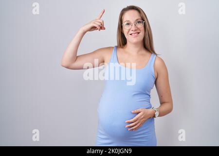 Giovane donna incinta in piedi su sfondo bianco sorridendo stupito e sorpreso e puntando in su con le dita e le braccia sollevate. Foto Stock