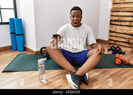 Giovane uomo africano seduto sul tappeto di allenamento in palestra con smartphone in faccia d'urto, con un aspetto scettico e sarcastico, sorpreso dalla bocca aperta Foto Stock