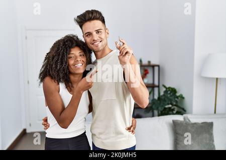 Giovane coppia interracial che tiene le chiavi della nuova sede sorridente felice che indica con la mano e il dito Foto Stock