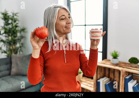 Donna di mezza età dai capelli grigi sorridente sicura che tiene la mela rossa e la dentiera a casa Foto Stock