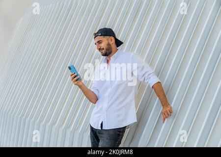 Giovane uomo ispanico in abiti casual e berretto a rovescio utilizzando il suo telefono cellulare appoggiato a una moderna struttura bianca slat. Foto Stock