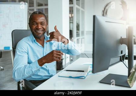 Uomo d'affari africano che usa il linguaggio dei segni mentre parla in linea con il cliente seduto in ufficio Foto Stock