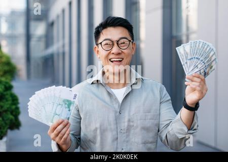 Felice ricco asiatico di mezza età che tiene due tifosi di soldi, celebrando la fortuna e il successo finanziario grande, in posa all'aperto Foto Stock