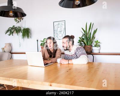 Maschi e femmine concentrati guardando lo schermo del computer portatile mentre si siede al tavolo di legno con tazze di caffè in appartamento Foto Stock