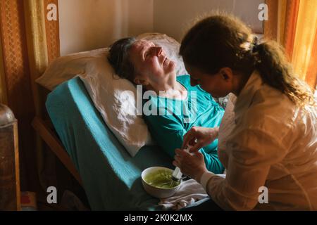 Infermiera che allatta la donna anziana paralizzata con purea attraverso il tubo e la siringa mentre si prende cura del paziente a casa Foto Stock