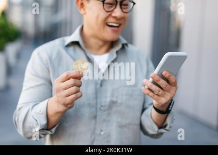Commercio in linea. Felice uomo asiatico in possesso di bitcoin dorato e smartphone, maschio di mezza età che guadagna criptovaluta Foto Stock