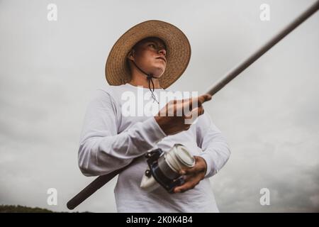 Da sotto maschio in cappello con canna da pesca cattura di pesce sulla riva sabbiosa Foto Stock
