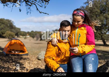 Giovane uomo seduto sulla roccia vicino a una tenda che abbraccia la ragazza in un outerwear che naviga sullo smartphone mentre accampano insieme in campagna Foto Stock