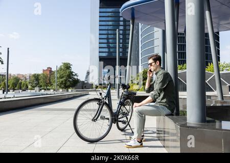 Vista laterale di un determinato maschio che effettua una chiamata telefonica sul cellulare mentre si siede vicino alla bicicletta in centro Foto Stock