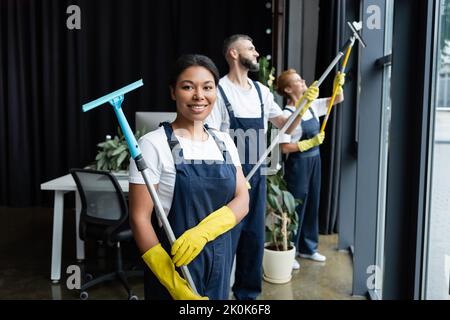 allegro misto gara donna che tiene tergicristallo vicino ai colleghi che lavorano in ufficio, immagine stock Foto Stock