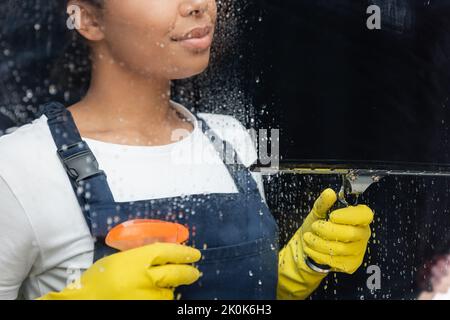 vista ritagliata della donna birazziale con detergente e finestra squeegee vicino al vetro bagnato in ufficio, immagine stock Foto Stock
