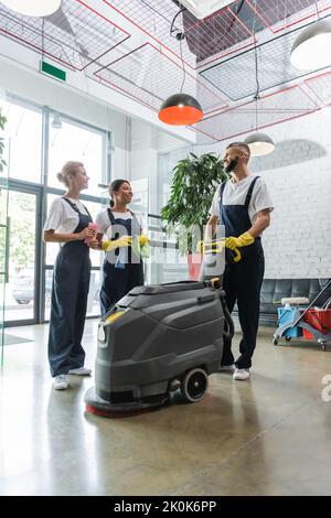 pulitori professionali multietnici felici che parlano vicino alla macchina lavapavimenti del pavimento nella lobby dell'ufficio, immagine di scorta Foto Stock