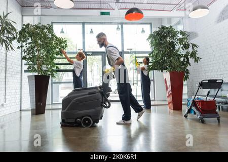 vista laterale dell'uomo in tute con lavapavimenti elettrici vicino a colleghi interracial, immagine stock Foto Stock