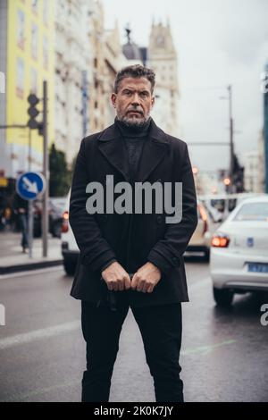 Uomo d'affari di mezza età in cappotto nero con ombrello e camminando sul marciapiede nelle giornate di pioggia Foto Stock