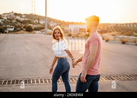 Vista laterale di una giovane coppia in abiti informali alla moda che camminano tenendo le mani sulla strada contro la città offuscata sulla collina durante il tramonto Foto Stock