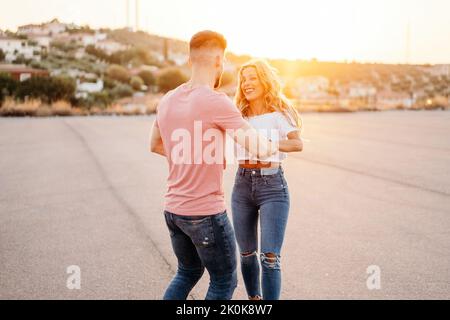 Uomo allegro e donna eccitata che si guardano e ridono mentre camminano danzando tenendo le mani lungo la strada asfaltata contro la città offuscata sulle colline Foto Stock