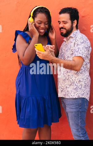 Felice donna nera in cuffie ascoltare la musica mentre si è in piedi vicino a un uomo etnico condividere playlist sullo smartphone contro la parete arancione in strada Foto Stock