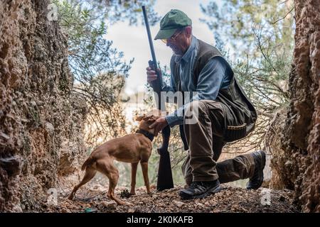 Vista laterale del cacciatore maschio in mimetizzazione e cappello in piedi sul ginocchio mentre accarezzano il cane da caccia tra le rocce in campagna Foto Stock