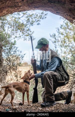 Vista laterale del cacciatore maschio in mimetizzazione e cappello in piedi sul ginocchio mentre accarezzano il cane da caccia tra le rocce in campagna Foto Stock