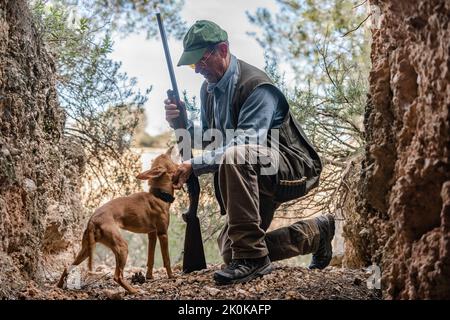 Vista laterale del cacciatore maschio in mimetizzazione e cappello in piedi sul ginocchio mentre accarezzano il cane da caccia tra le rocce in campagna Foto Stock